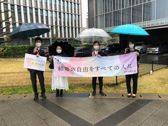 裁判所の前で、「結婚の自由をすべての人に」と書かれた九州訴訟特製の横断幕やプラカードを持つ４人の人達。雨が少し降っていて、みんな傘をさしている。