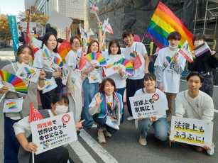 パレードに参加した北九州市立大学の学生さん１１人と先生の集合写真
マリフォーのYESの旗や、法律上も家族にや法律上もふうふにや同性婚で世界はもっとしあわせになる、社会は変わってしまわないなどと書かれたボードを持ったり、レインボーのアイテムを持ったりしてしている。