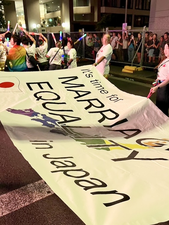 パレードを歩くひとたちの姿。
レインボーに光る棒を持って踊っているひとたちと、大きな布を広げているひとたちがいる。

大きな布には、「it's　time for MARRIAGE WQUALITY in Japan」（日本でも婚姻平等を実現する時だ）というメッセージが書かれている。
