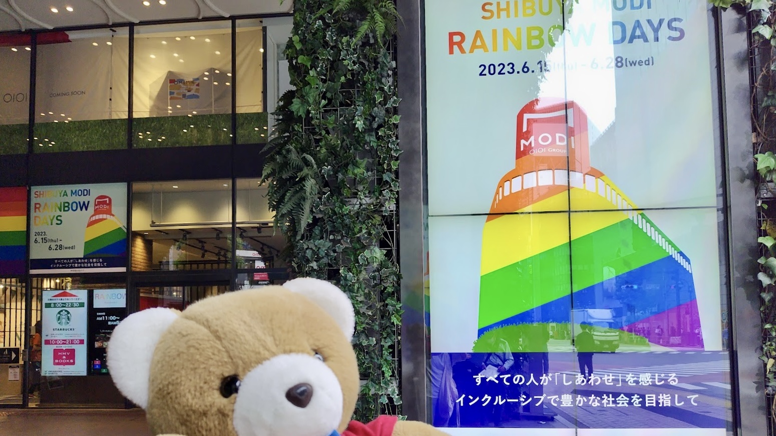 商業施設「渋谷MODI」の前でくまが手を上げてポーズをとっている。 MODIには、大きな電光掲示版が複数設置されていて、レインボーカラーになっていたり、「RAINBOW DAYS　すべての人が「しあわせ」を感じるインクルーシブで豊かな社会を目指して」というメッセージが表示されている。