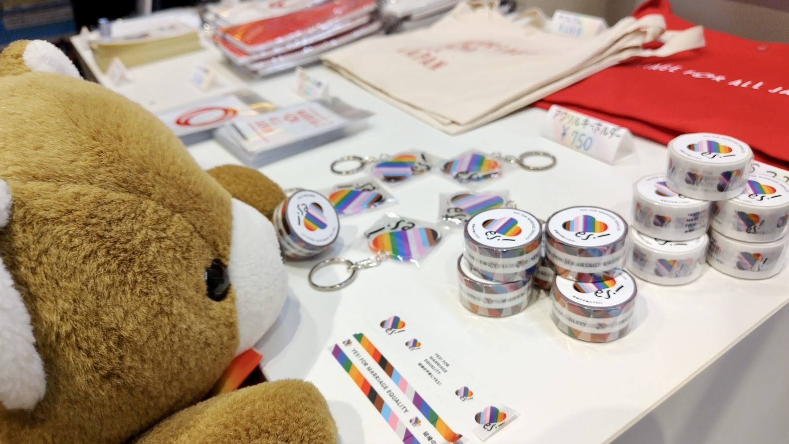 「結婚の平等にYES！」と書かれたマスキングテープや、YES！のロゴのキーホルダーが飾られている。 奥には、「MARRIAGE　FOR　ALL　JAPAN」と書かれたサコッシュも並んでいる。 クマは、マスキングテープを手に取り、珍しそうに眺めている。