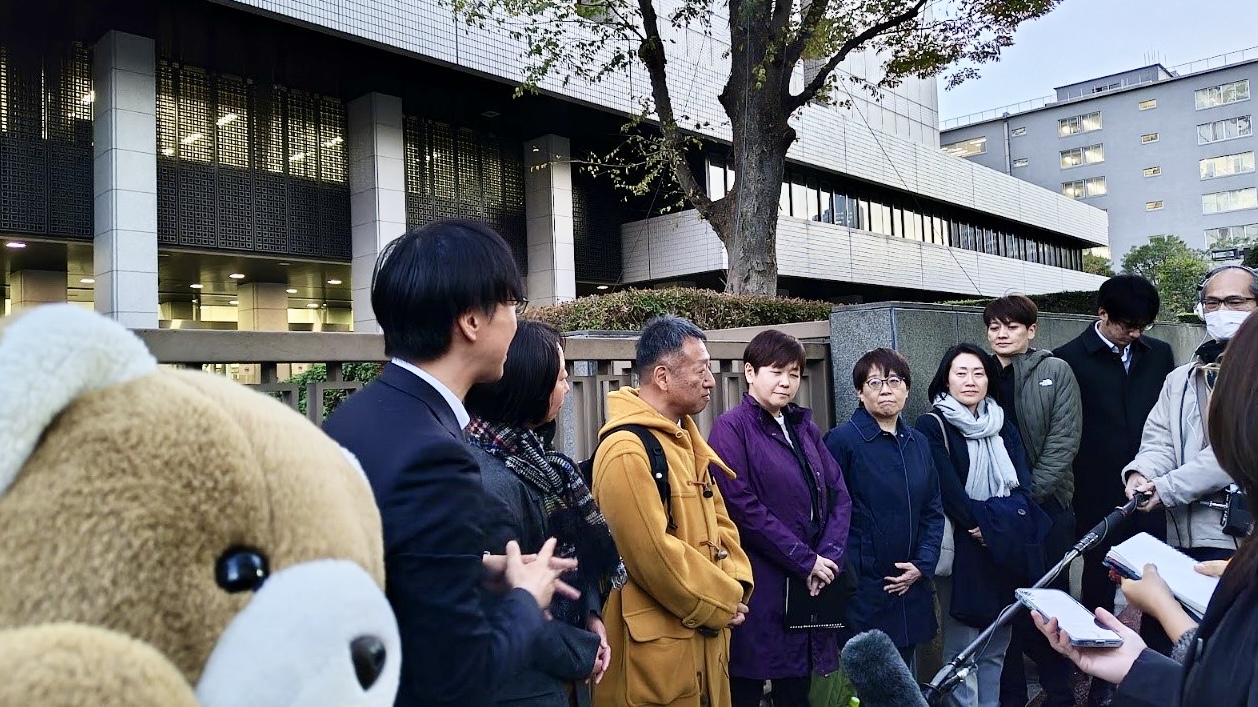 東京地裁前に、結婚の自由をすべての人に東京２次訴訟の原告・弁護士が並んでいる。 その前には、カメラやレコーダーをもった記者がいる。 記者たちが原告さんに取材をして、原告さんが返答している。 くまはがんばれーという顔で取材風景を見守っている。