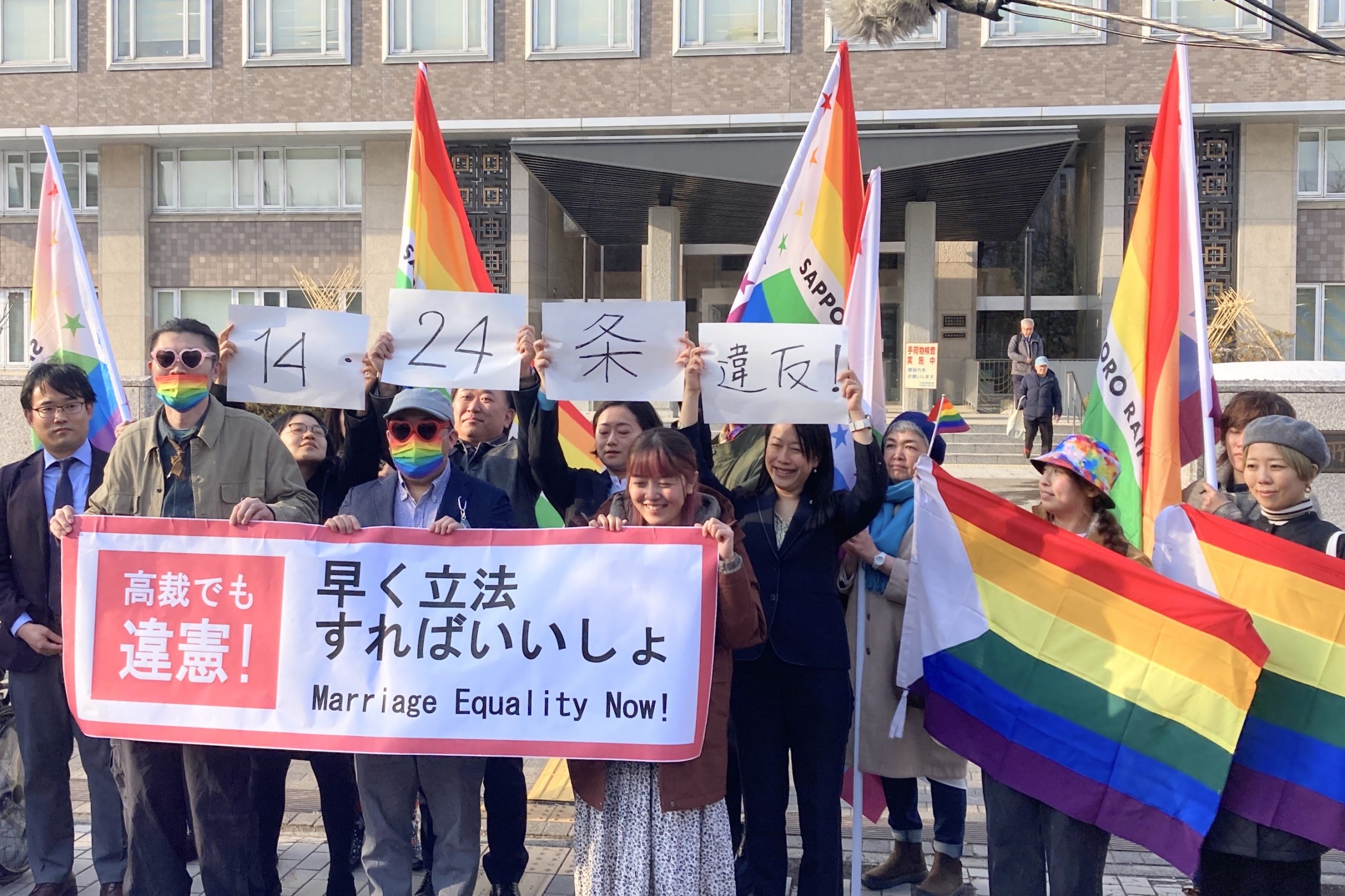 札幌地方裁判所の前で、「結婚の自由をすべての人に」北海道訴訟の原告と弁護団、支援者が、「高裁でも違憲！早く立法すればいいしょ」と書かれた横断幕やレインボーカラーの旗を掲げている