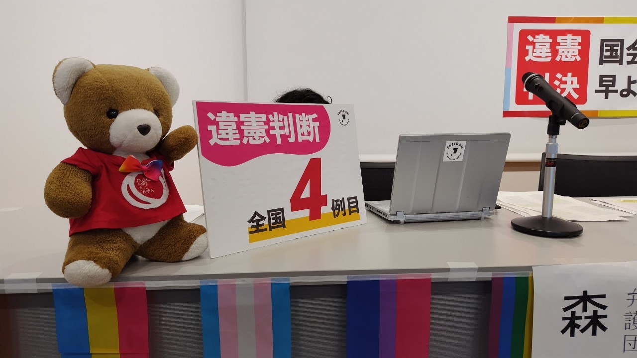 報告会の壇上の様子 マリフォーのTシャツを着たクマのぬいぐるみの側に違憲判断全国４例目と書かれたボードが飾られている