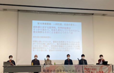 弁護士２名と原告さん４名が壇上に座っている。原告提出書類の内容を説明するスライドが映し出されている。