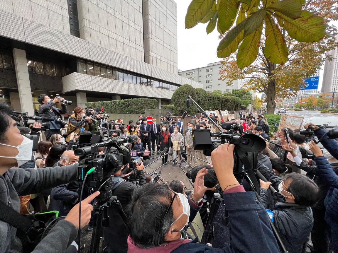 ③ 判決言渡し前に裁判所前で多数の報道陣に囲まれて取材を受ける原告らの様子。