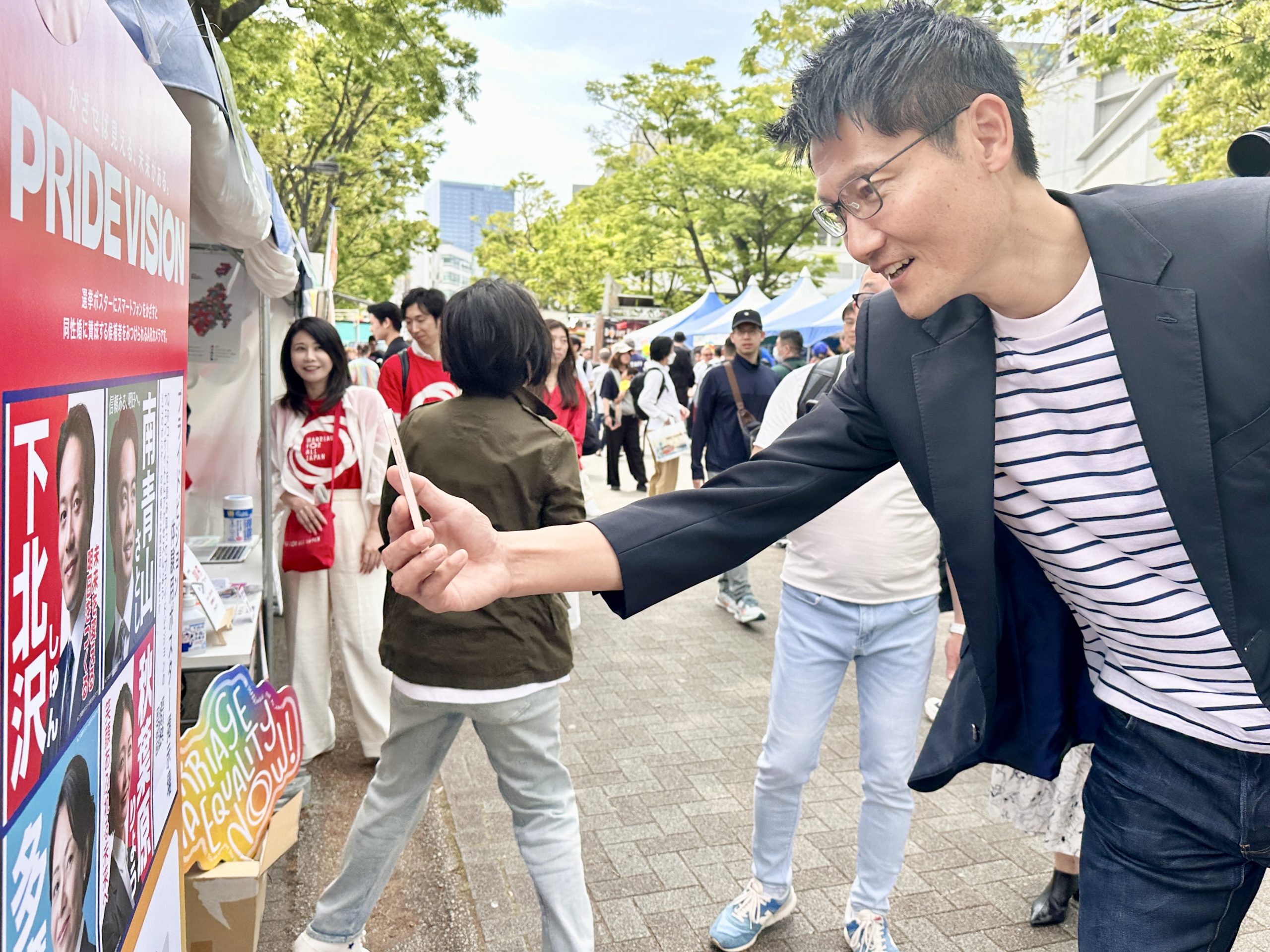 東京レインボープライド2024のマリフォーブース前にて、選挙ポスターにスマートフォンをかざすと、結婚の平等（同性婚）法制化に賛成の候補者を見分けられるARカメラ「PRIDE VISION（プライドビジョン）」を体験する、自民党の朝日けんたろう参議院議員