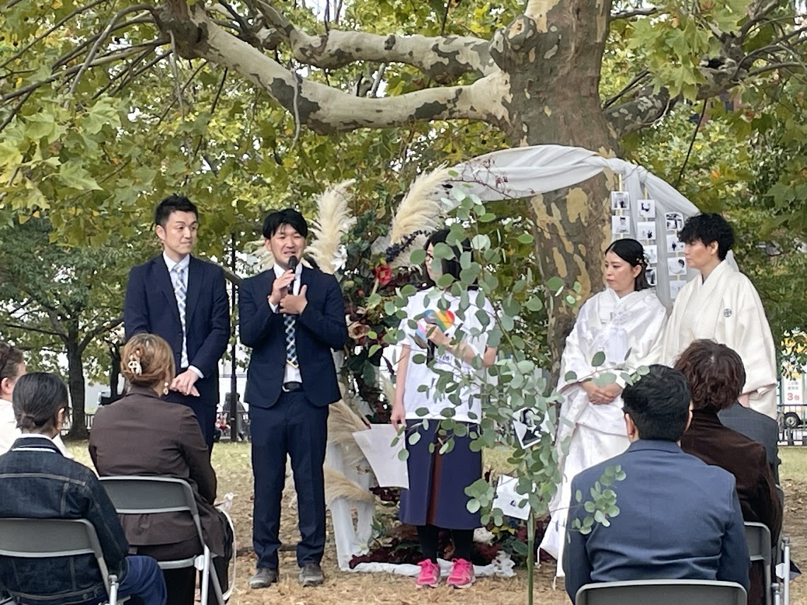 結婚式の写真 外で行われた。木の下に結婚式をあげる女性どうしのカップルがおり、そばに司会をしている男性どうしのカップルがいる。前には参列者の人たちが座っている。カップルの間に、弁護団員がいるが、木に隠れてあまり見えない。