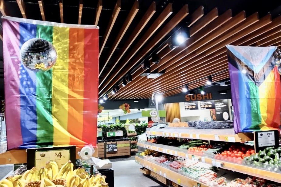 オーストラリア・シドニーのスーパーマーケットの野菜・果物売り場。 棚にさまざまな野菜や果物が並べられている。 天井からは、大きなレインボーフラッグとミラーボールがつるされている。