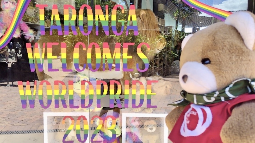 動物園のお土産やさんをくまがのぞいている。 お土産さんのガラスには、「TRONGA WELCOMES WORLDPRIDE 2023」（タロンガ動物園は２０２３年ワールドプライドを歓迎します）というメッセージが書かれている。
