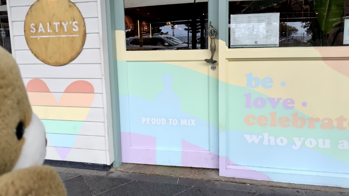 くまがカフェに入ろうとしている。 カフェの扉には、レインボーカラーのハートマークや、「PROUD TO MIX」「be.love.celebrate. who you are」（あなたが自身でいて、あなた自身を愛して、あなた自身を祝福して）というメッセージが描かれている。
