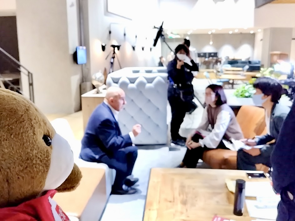 ソファーに腰かけて話している人たちの様子を くまが見守っている。 話している人の一人はEvanさん。 アナウンサーと通訳さん、撮影スタッフがEvanさんを囲んで取材している。