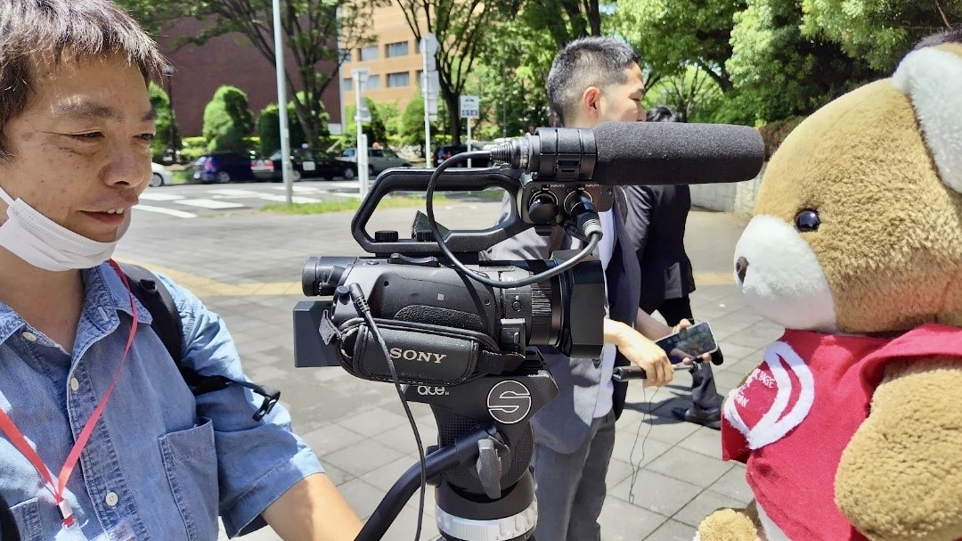 裁判所前の路上。 カメラを構えた人が笑っている。 カメラのレンズの先には、くまがいる。 その奥にも、打合せをしているひとたちがいる。