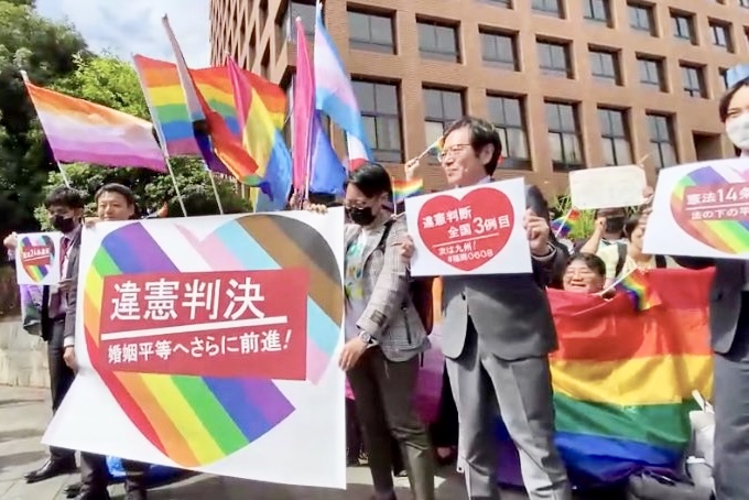 名古屋地方裁判所の前にたくさんの人が並んでいる。笑って喜んでいる人や、感極まって泣いている人もいる。 たくさんのレインボーフラッグがはためく中、大きく「違憲判決 婚姻平等へさらに前進！」と書かれたプログレスフラッグカラーの旗が広げられている。 その横には、「違憲判断全国３例目」と書かれたメッセージをもっている人がいる。