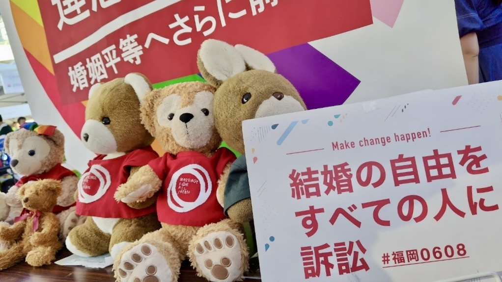 マリフォーのロゴ入りのシャツをきたくまのぬいぐるみが並んでいる。うさぎのぬいぐるみもいる。 うさぎは、「結婚の自由をすべての人に訴訟 ＃福岡０６０８」と書かれたパネルを持っている。 くまとうさぎの奥には、「違憲判決 婚姻平等へさらに前進」と書かれた大きな旗が飾られている。