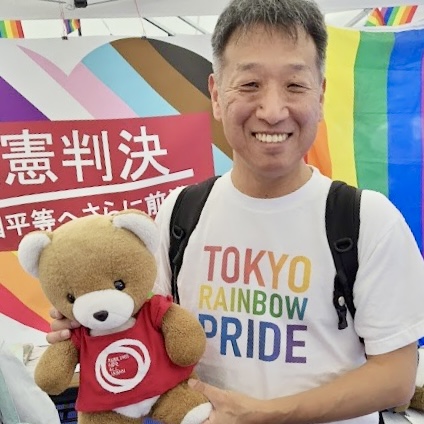 「違憲判決 婚姻平等へさらに前進」と書かれた旗の前で、男性がくまのぬいぐるみを抱いている。 男性は、東京２次訴訟原告の山縣さんで、「TOKYO RAINBOW PRIDE」と書かれたシャツを着ている。 くまは、マリフォーのロゴが入ったシャツをきている。