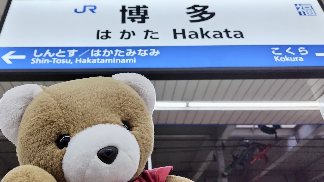 博多駅のホームで、「博多」という看板の前でポーズをとるくま