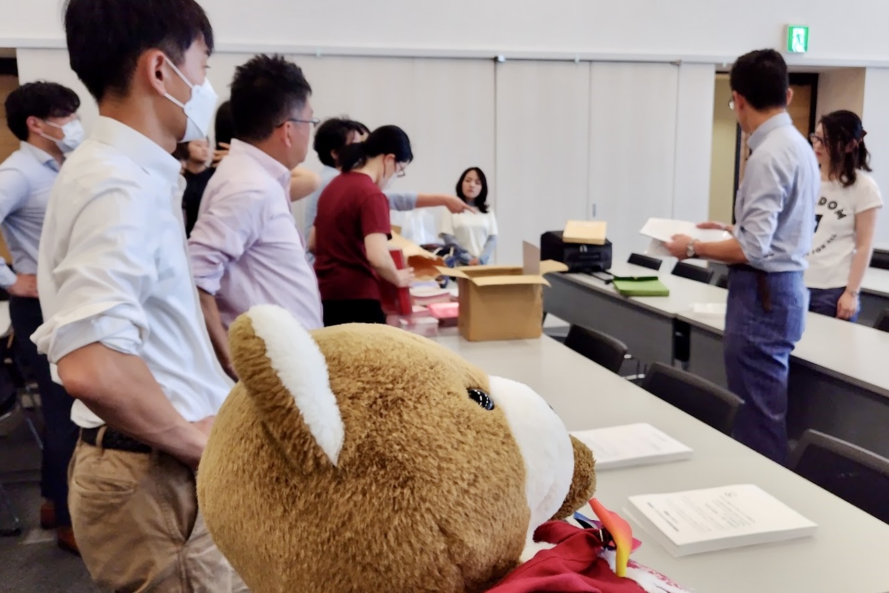 広い会議室で、大勢の人が書類を並べたり話をして報告会の準備をしている。 くまはがんばれーと応援している。