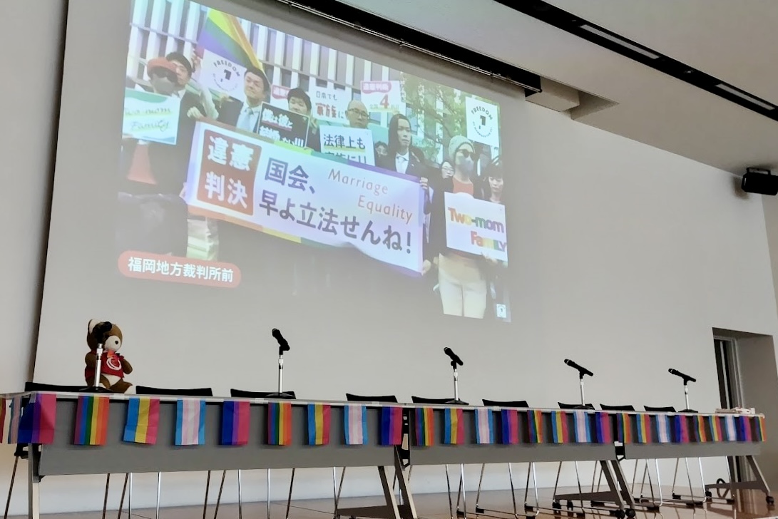 大きな会議室の前方に、福岡地裁前の様子が生中継で写っている。 福岡地裁前では、九州訴訟の原告さんと弁護団がならんでいて、「違憲判決 国会、はよ立法せんね！」と書かれた旗を広げ、意見判決が出たことを知らせている。 中継画面の手前には、くまが座っている。