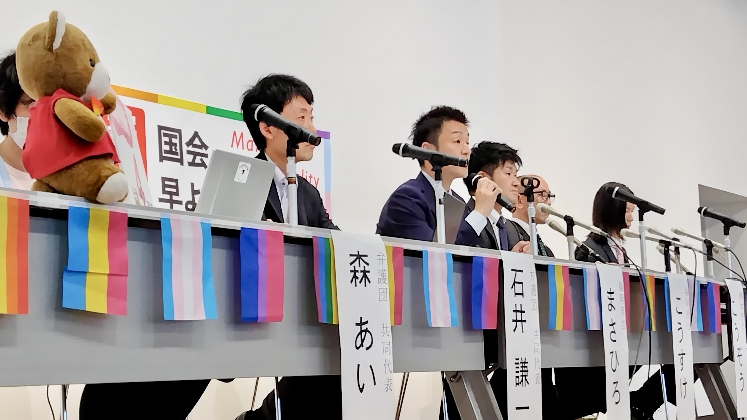 会議室前方に、九州訴訟の弁護団、原告さんが並んでいる。 弁護団と原告さんの前にのテーブルには、マイクがずらりと並んでいる。 原告まさひろさんがマイクをもち、判決の感想を話している。