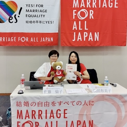 マリフォー国会司会席の様子。 壁の手前にテーブルがあり、二人の弁護士が座っている。 テーブルに「佐藤倫子」「須田布美子」の名札が置かれている。 マリフォーの横断幕がテーブルや壁に飾られている。 佐藤弁護士が、くまを抱き、須田弁護士がマリフォーのチラシを掲げ、肩を寄せてポーズをとっている。