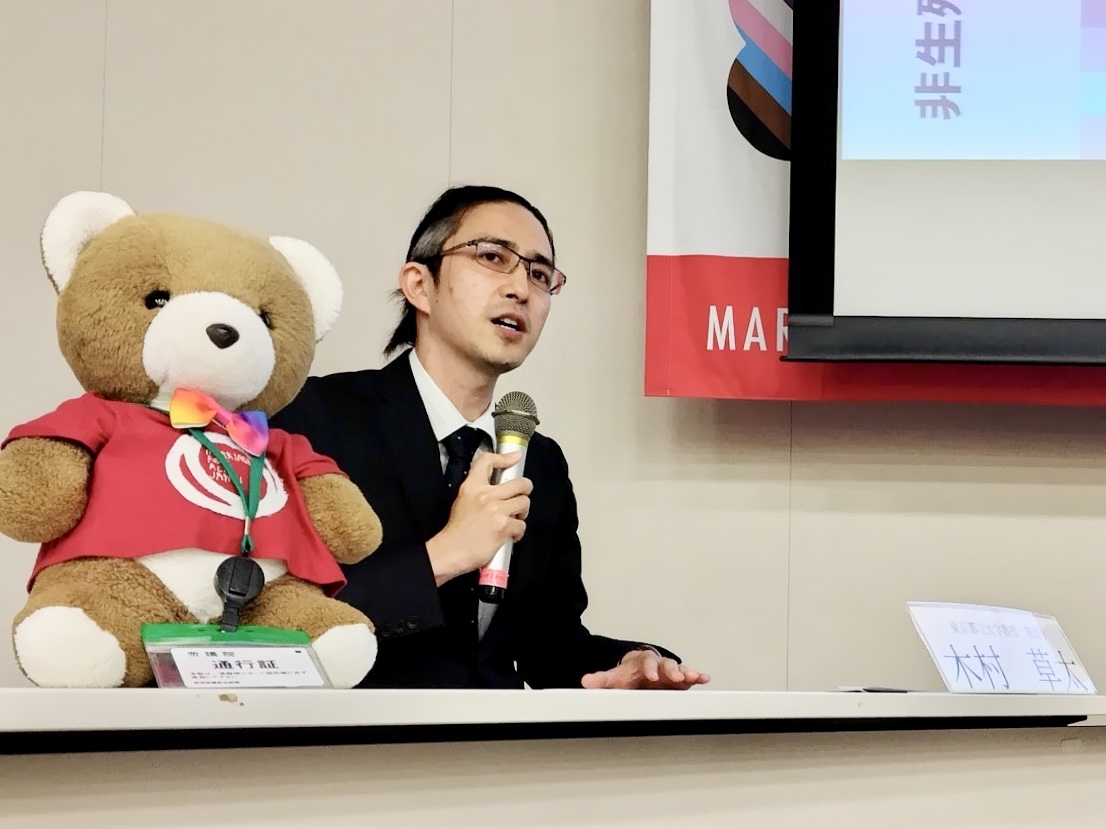 マリフォー国会司会席のそばのテーブルに、憲法学者の木村草太さんが座っている。 マイクを持って話をしている。 木村さんのそばにくまがいる。 司会席から佐藤弁護が真剣な目で話を聞いている。