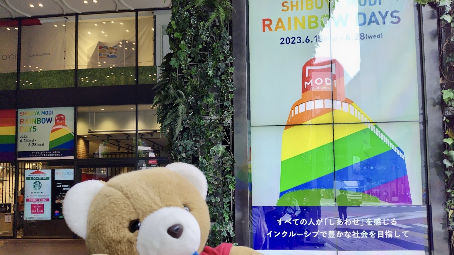 商業施設「渋谷MODI」の前でくまが手を上げてポーズをとっている。 MODIには、大きな電光掲示版が複数設置されていて、レインボーカラーになっていたり、「RAINBOW DAYS すべての人が「しあわせ」を感じるインクルーシブで豊かな社会を目指して」というメッセージが表示されている。