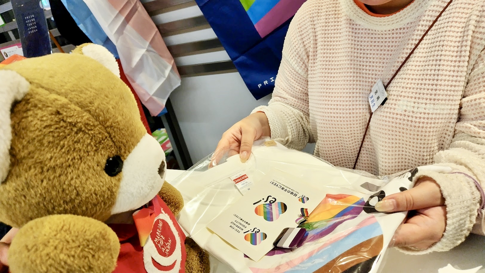 くまがレジで店員さんから品物を受け取っている様子。 品物は、「結婚の平等にYES」のロゴが入ったシャツやステッカー、ノンバイナリーのピンバッジ、アセクシュアルのピンバッジ。