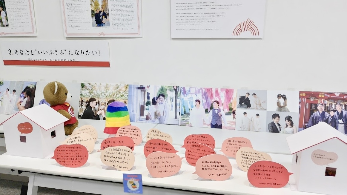 壁に、法律上同性のカップルたちの写真がたくさん飾られている。 その手前のテーブルには、カップルたちのメッセージパネルが飾られている。 メッセージパネルの隙間に、くまとぶらんちゃんが向き合って座って、おしゃべりしている。