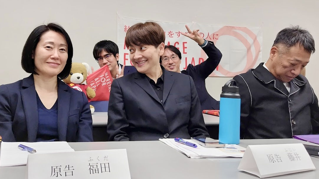 尋問期日報告会に登壇した東京２次原告の福田さん、藤井さん、山縣さん、三浦弁護士、沢崎弁護士、くま。 福田さんと藤井さんはカメラに気づいて笑顔を見せている。山縣さんはカメラに気づかず下を向いている。 三浦弁護士と沢崎弁護士は楽しそうにポーズをとっている。