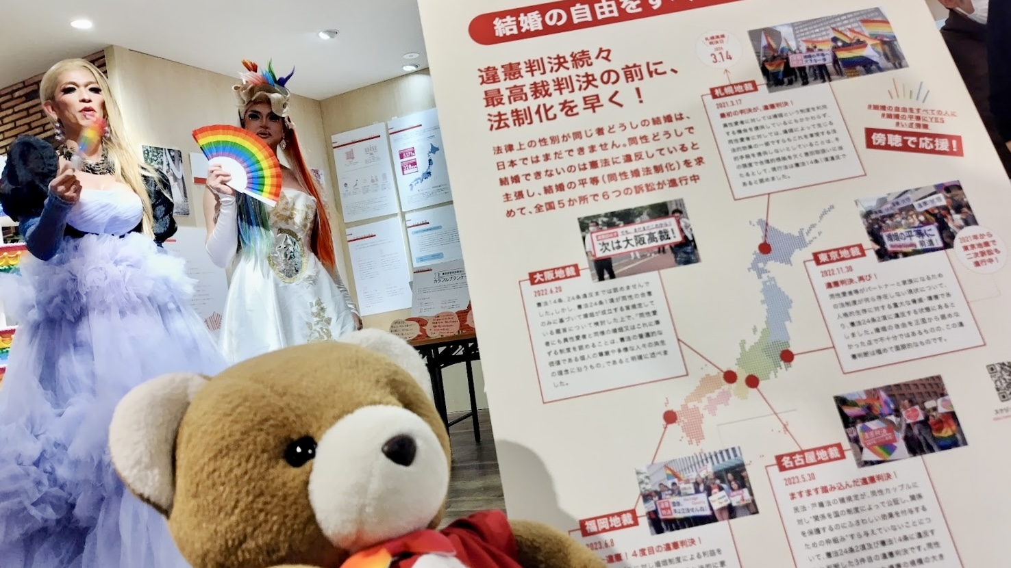 「結婚の自由をすべての人に訴訟 違憲判決続々 最高裁判決の前に、法制化を早く！」というパネルの前で、くまが目を輝かせている。 パネルには、結婚の自由をすべての人に訴訟の５地裁判決それぞれのポイントが記載されていて、「傍聴で応援」という呼びかけられている。 くまの奥には、お姫様のようなドレスをきたドラァグクイーンが二人経っていて、くまを見守っている。
