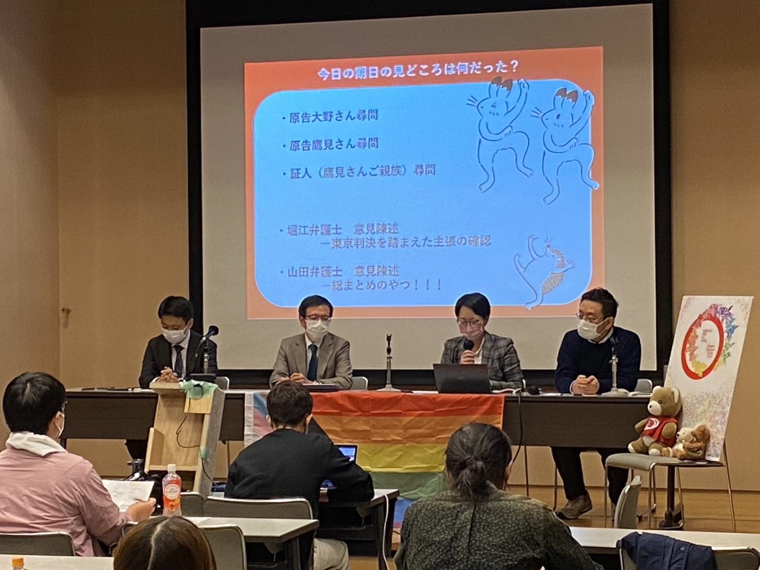 （期日報告会の様子。檀上に弁護団３名と、支援者の風間孝さんが並んでいる。マリフォーシャツを着たクマと原告カップルを模したクマたちも登壇者の隣で一緒に並んでいる。）