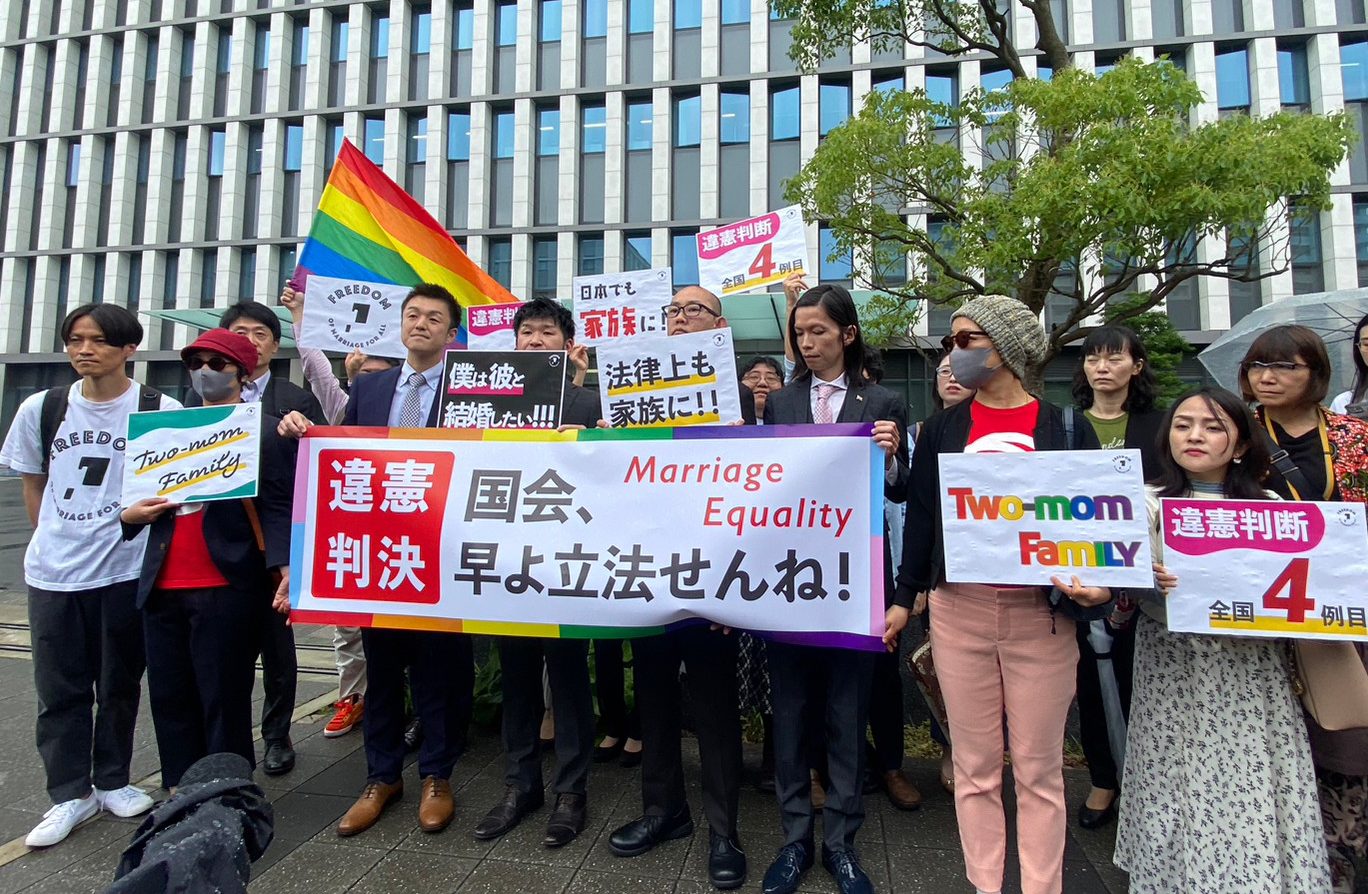 裁判所前で、判決内容を報告する旗を出す原告と弁護団 原告が持っている旗には、違憲判決、国会、早よ立法せんね marriage equality などと書かれている。 他にも、two mam family 、法律上も家族に、僕は彼と結婚したい 日本でも家族に、違憲判断全国４例目などと書かれている。