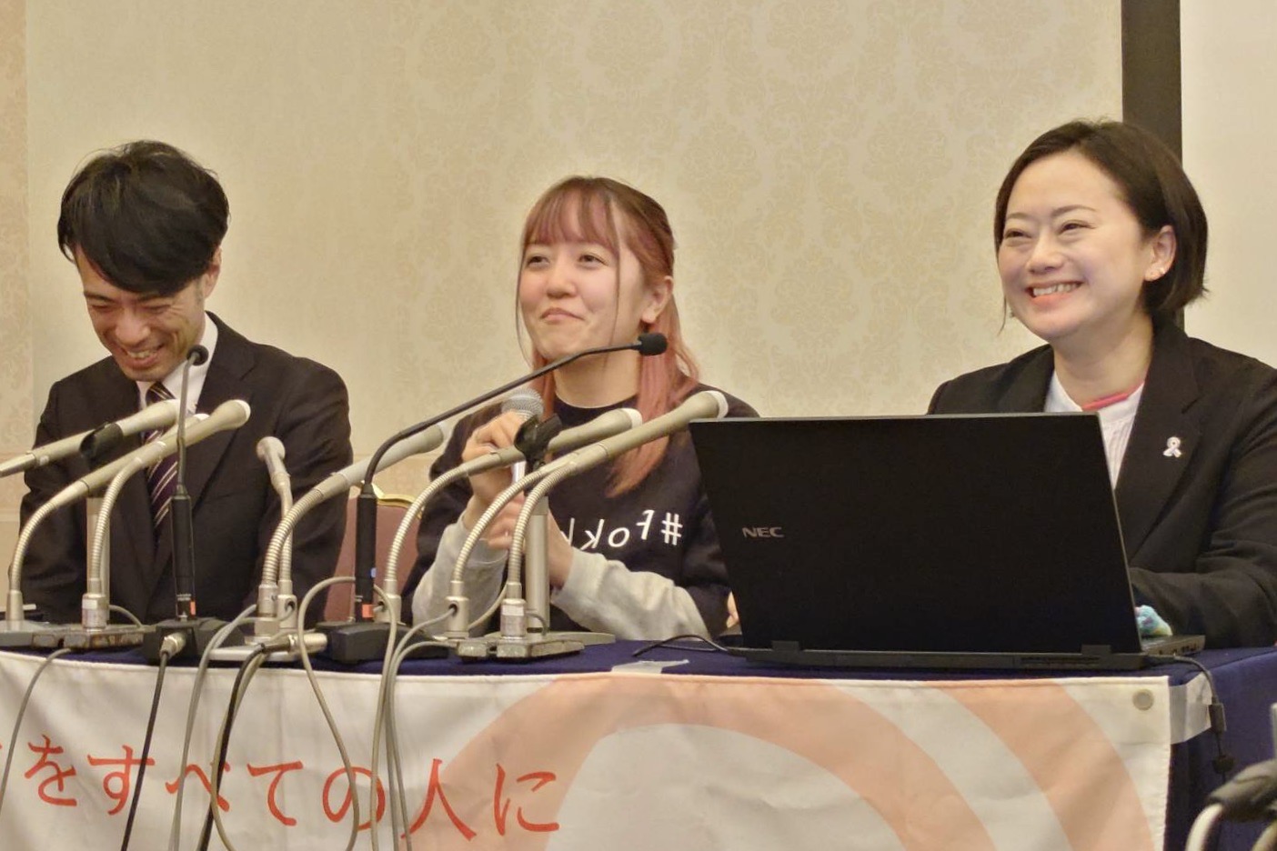 記者会見の場で、原告の中谷衣里さんが笑顔を見せている。中谷衣里さんの左側には綱森史泰弁護士が、右側には皆川洋美弁護士が座っている。