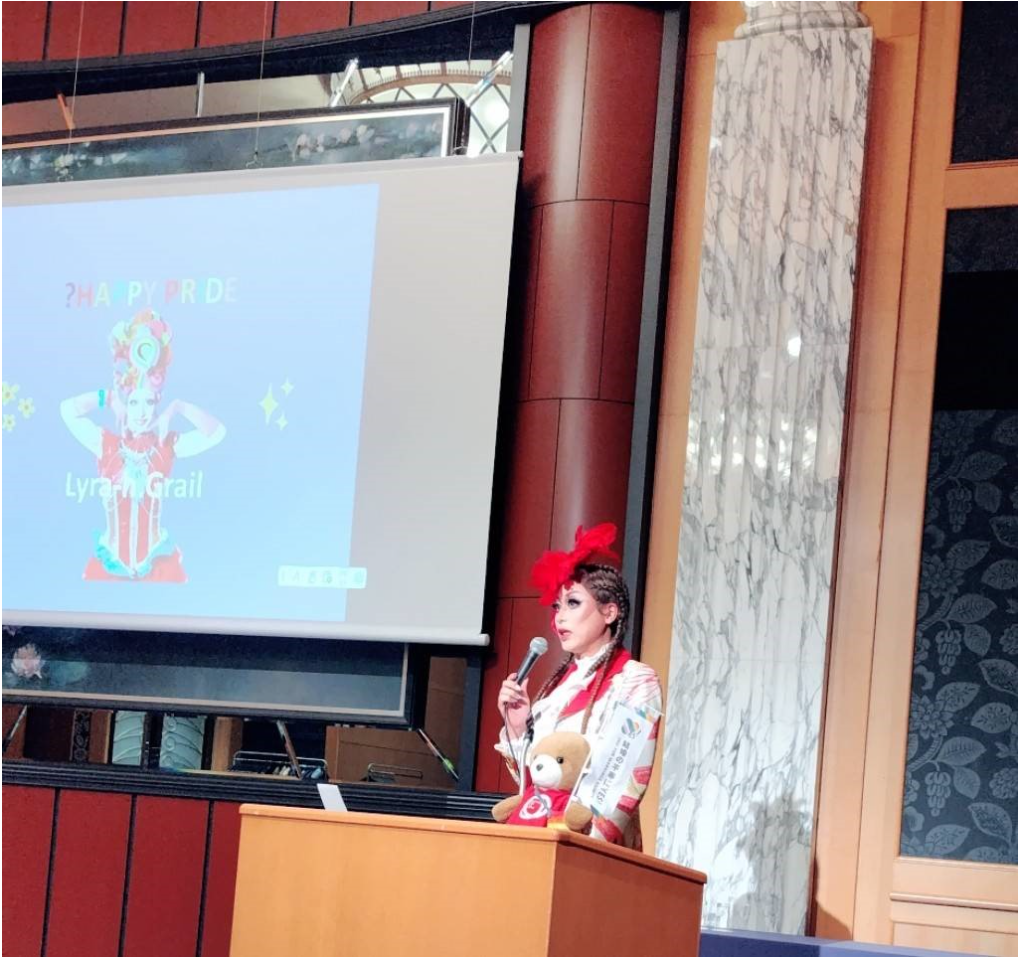 スクリーンにドラァグクイーンのライラの写真と「HAPPY PRIDE」という文字が表示されている。 演題には、ライラが立ってマイクを持って話をしている。ライラのそばにくまちゃんがいる。