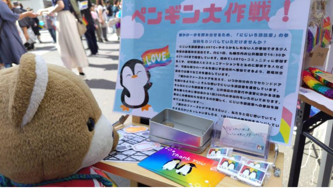 L-portブースに設置された「ペンギン大作戦」というタイトルのパネル。 性的マイノリティやそうかもしれない人が集まれる「にじいろ談話室」という活動をしていという紹介と、参加費２００円がハードルになって参加しづらい人のために参加費を寄付してほしいというお願いが書かれている。 くまちゃんが、すてきな取り組みだなあと感心しながらパネルを眺めている。