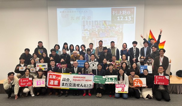 報告会後の集合写真 原告、弁護団、支援者、マリフォーのスタッフで並んで、横断幕などを持って笑顔で写っている。