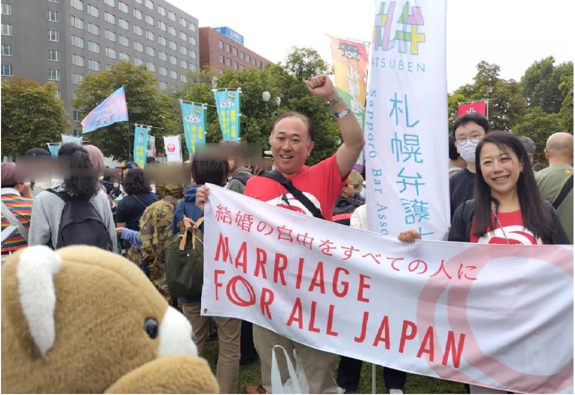 パレード出発地点の写真。 たくさんの人がパレードのために整列している。 加藤弁護士と須田弁護士が「結婚の自由をすべての人に」と書かれた横断幕をもって、笑っている。 その奥には、札幌弁護士会の旗をもった人が立っている。