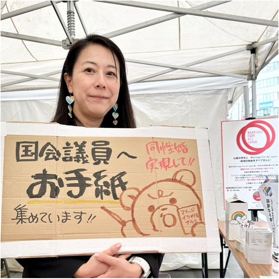 マリフォーブース内の写真。 須田弁護士がパネルを持って、笑顔で立っている。 パネルは段ボール製で、手書きで、「国会議員へお手紙を集めています」と書かれている。 怒り顔のくまちゃんが「同性婚実現して」と言いながらお手紙を書いているイラストが添えられている。
