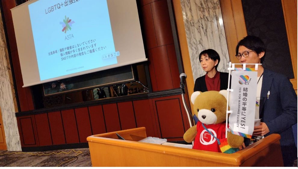 披露宴のスクリーンが設置され、「LGBTQ＋出張授業」と表示されている。 演題にASTAのスタッフが２名立っている。 演題の上に、くまちゃんがおり、くまはマリフォ―ロゴ入りのシャツを着て、「結婚の平等にYES」と書かれた旗を掲げている。