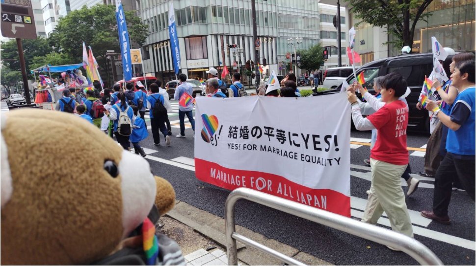 パレードの写真。結婚の平等にYES！と書かれた大きな幕を数名で掲げて歩いている。 その姿を沿道からくまちゃんが見ている。