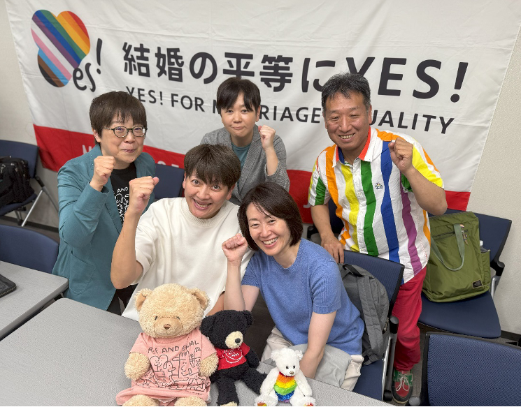 「結婚の平等にYES!」と書かれた横断幕の前で、笑顔でガッツポーズをとる「結婚の自由をすべての人に」東京２次訴訟の原告５人（後列は左から鳩貝さん、河智さん、前列は左から藤井さん、福田さん、山縣さん）。その前にはクマのぬいぐるみ３体が置かれている。