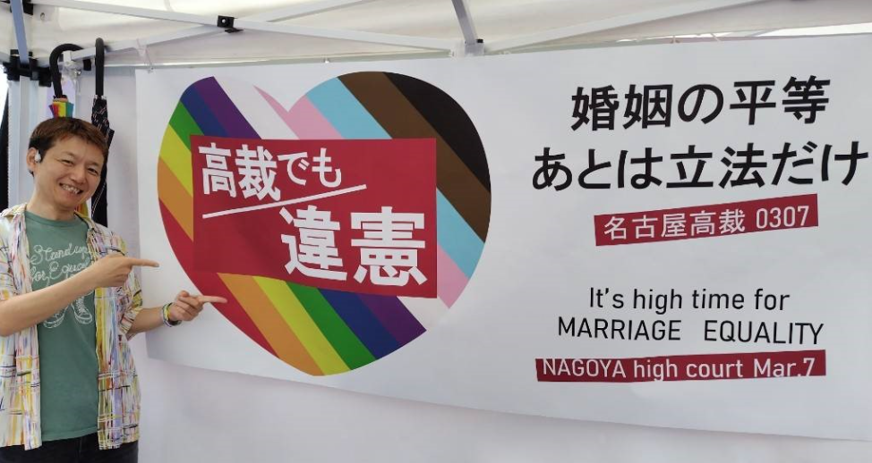 「高裁でも違憲　婚姻の平等あとはりっぽうだけ　名古屋高裁0307」と書かれた旗が壁に飾られている。その旗の隣で堀江弁護士が微笑んでいる。