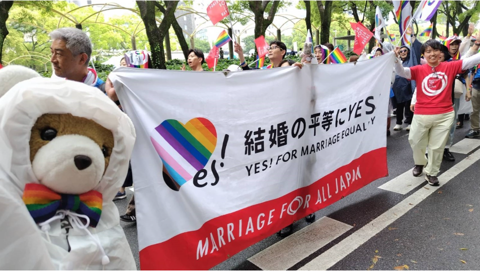 パレードの写真。 「結婚の平等にYES！」と書かれた幕を持った人たちの前で、くまちゃんがレインコートを着ている。 マリフォ―のシャツを着たスタッフが笑顔を向けている。