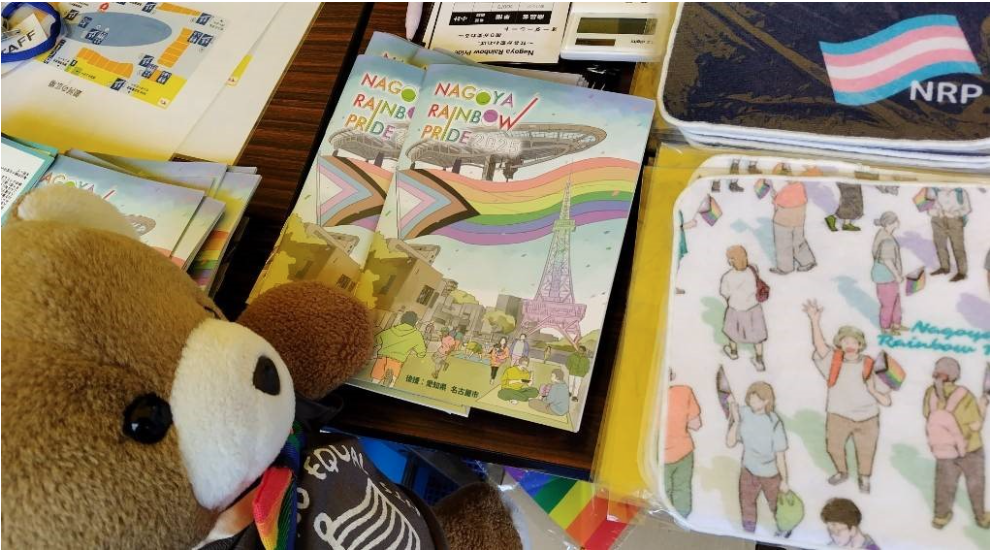 名古屋レインボープライドのパンフレットと公式グッズのハンドタオルが置かれたテーブルの写真。テーブル手前にくまちゃんがいる。
