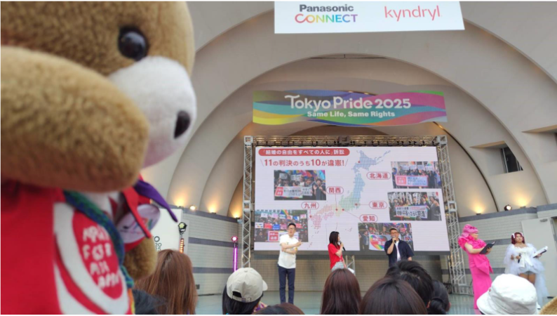 イベント会場内のステージを、観客席から眺めるくまちゃん。 ステージ上には、５名が立って話をしている。 ステージ上のスクリーンには、結婚の自由をすえての人に訴訟の判決を紹介する画像が映し出されている。