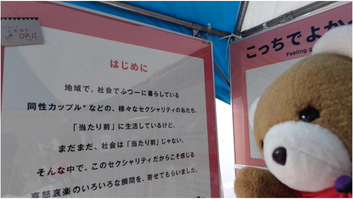 展示パネルを眺めるくまちゃん。 パネルには以下の説明が記載されている。 「はじめに　地域で、社会でふつーに暮らしている同性カップルなどの、様々なセクシュアリティの私たち。当たり前に生活しているけど、まだまだ、社会は当たり前じゃない。そんな中で、このセクシュアリティだからこそ感じる喜怒哀楽のいろいろな瞬間を、寄せてもらいました」