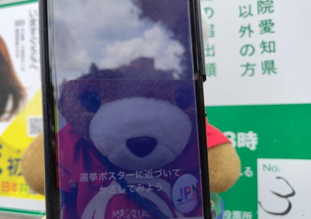 スマホのカメラが起動していて、「選挙ポスターに近づいてかざしてみよう」という文字が画面上に表示されている。 カメラはくまちゃんの姿をうつしている。