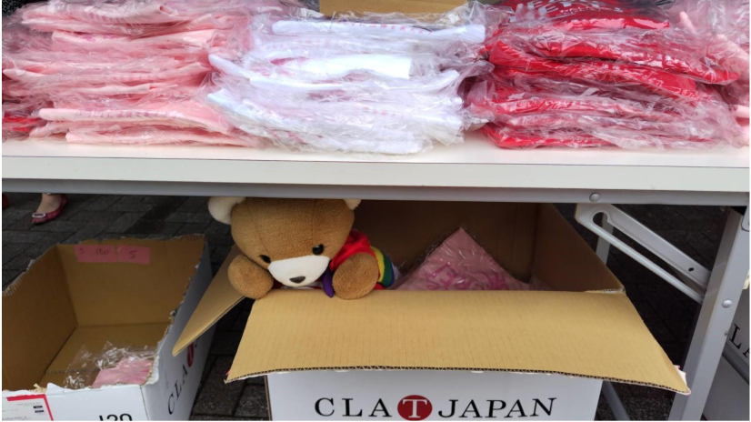 机の上にTシャツがたくさん並んでいる。 机の下にはシャツの入った段ボールが置かれ、くまちゃんが段ボールの中で手伝いをしている。