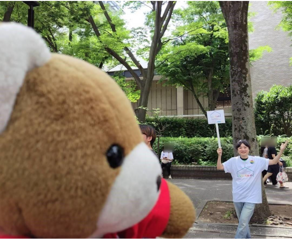 西川さんが「にじいろかぞく」とかかれた看板をもってポーズをとっている。くまが西川さんに手を振っている。